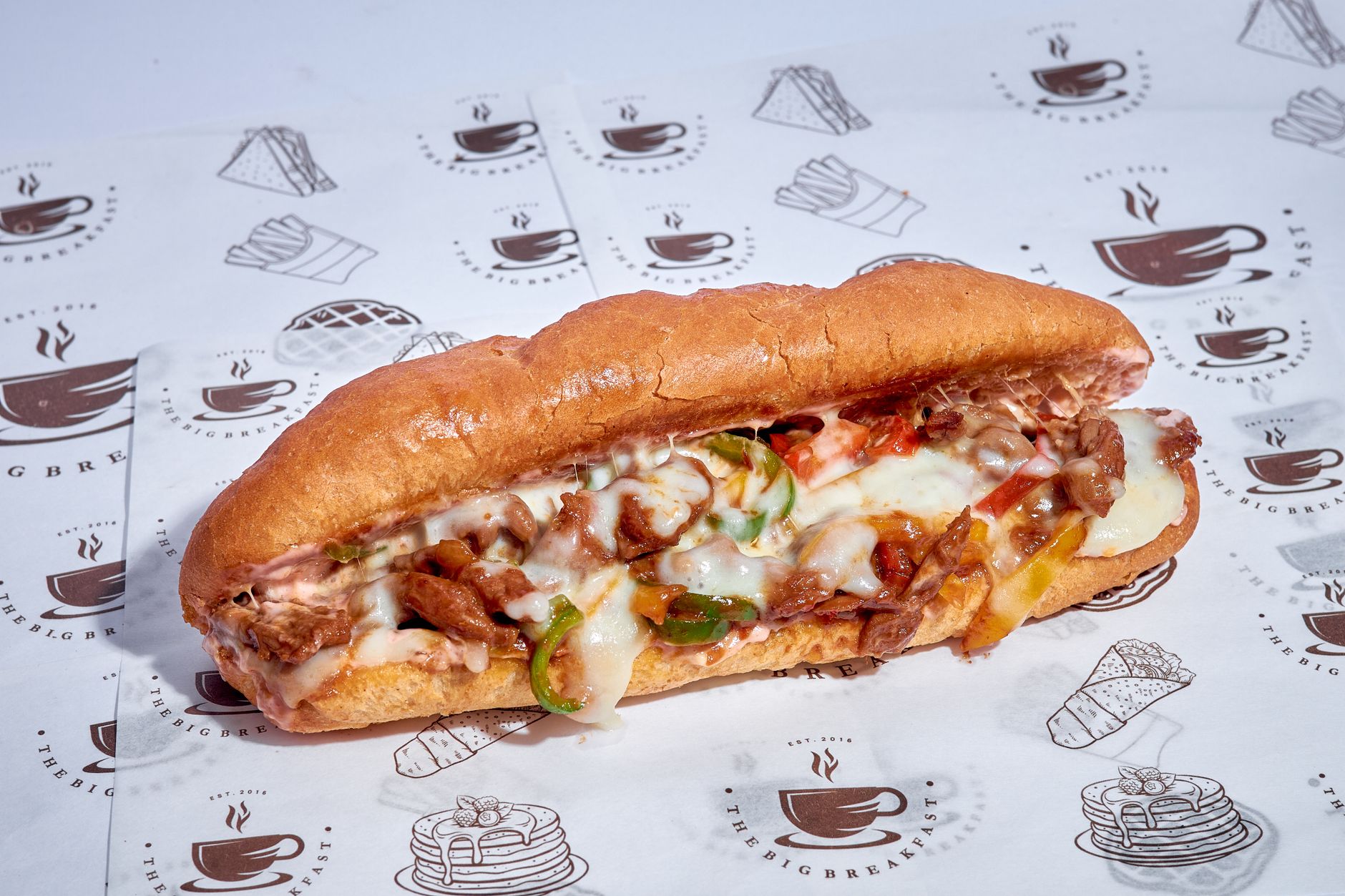 Close-up of a sub sandwich with melted cheese, peppers, and meat on branded paper.