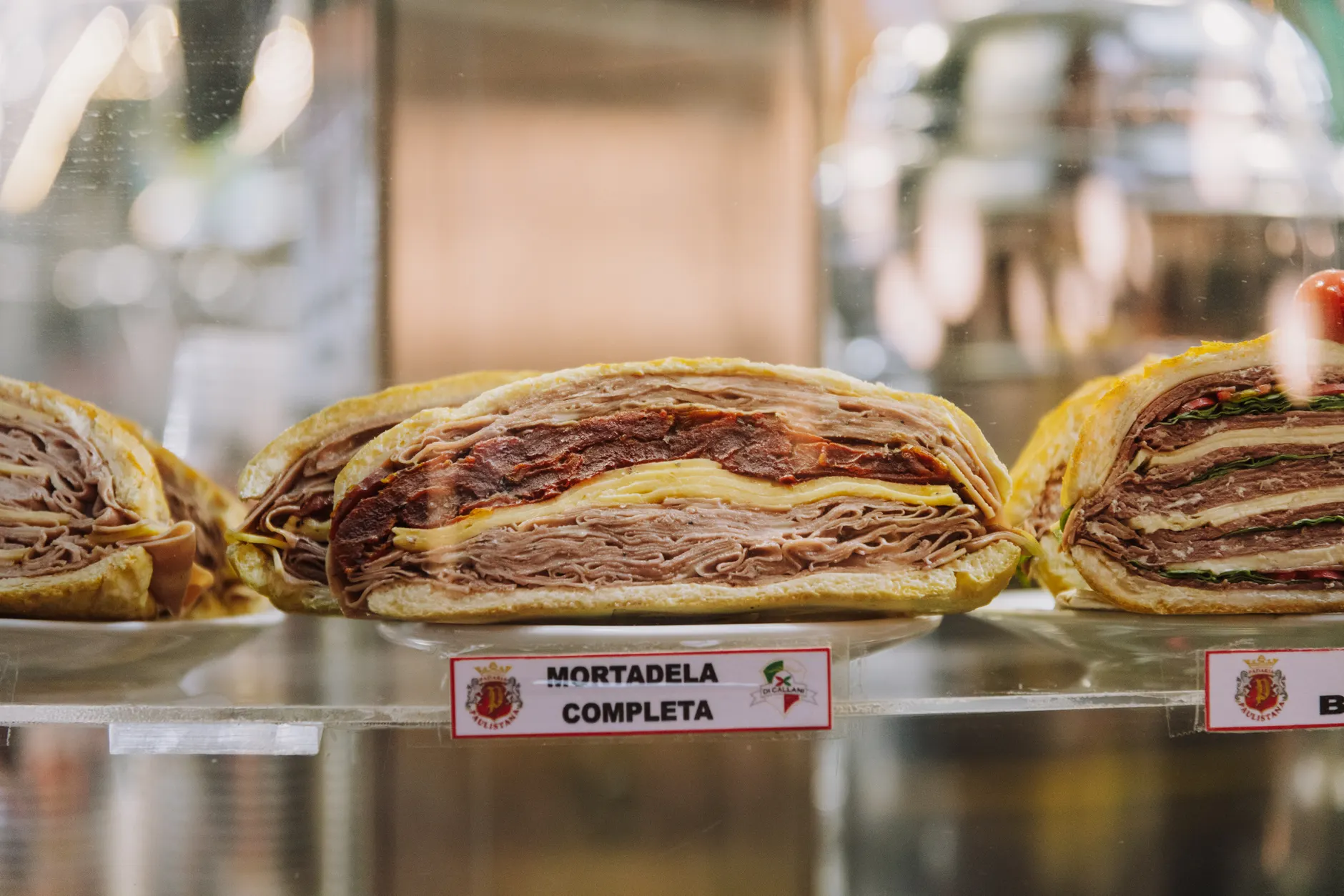 Assorted sandwiches with mortadela, cheese, and greens on display at a deli counter.